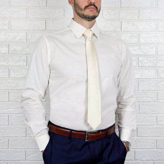 An individual in an ARTURO Slim Fit Long Sleeve Ivory Dress Shirt and a cream tie stands against a light brick wall, wearing navy pants with a hint of spandex and a brown belt.
