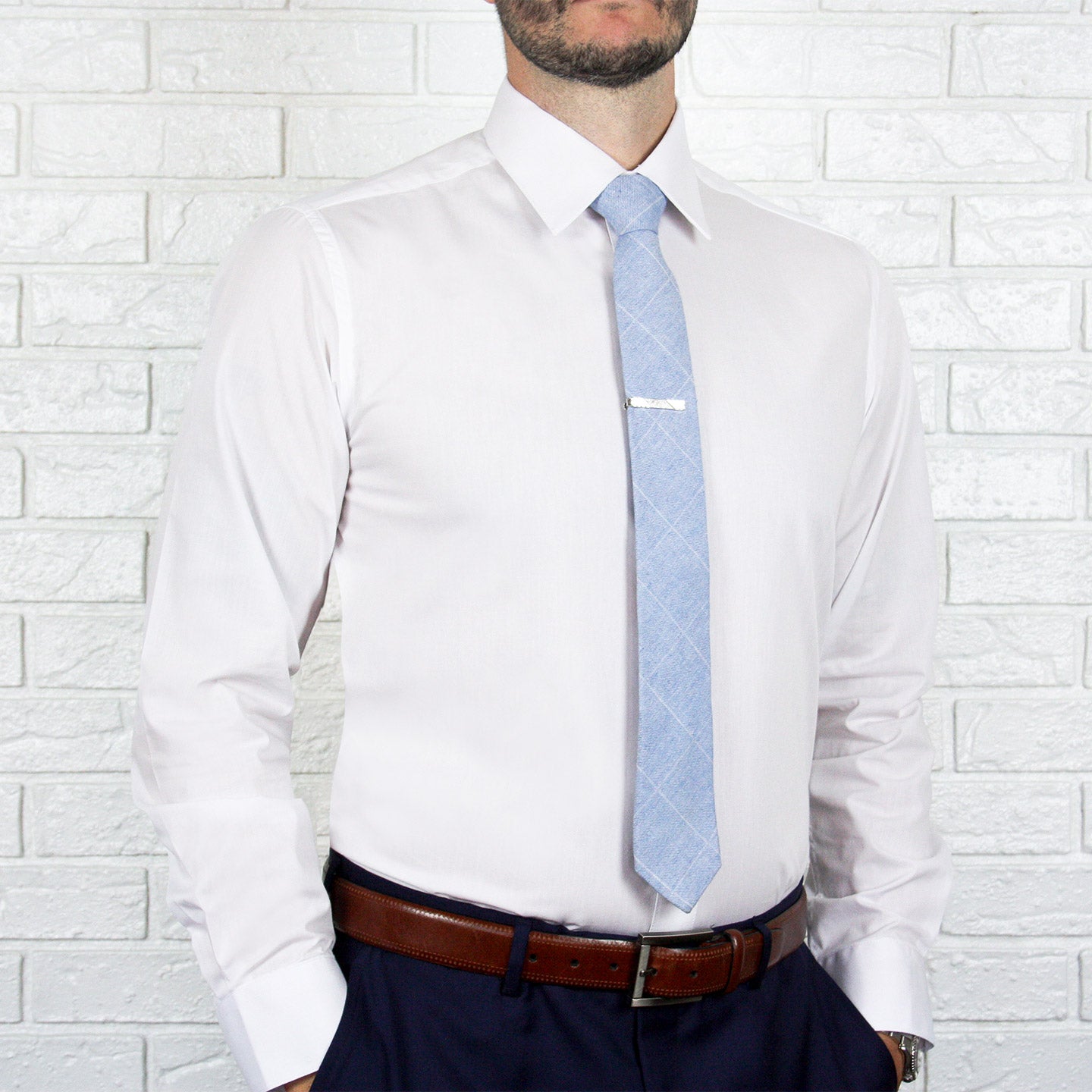 An individual stands against a white brick wall, showcasing an ARTURO Slim Fit Long Sleeve White Dress Shirt, beautifully complemented by a light blue tie with a sleek tie clip. His outfit is finished with a brown belt and dark pants, exuding style and sophistication.