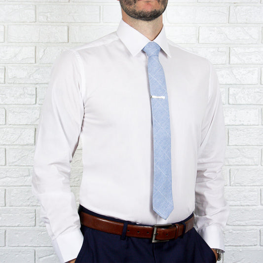 An individual stands against a white brick wall, showcasing an ARTURO Slim Fit Long Sleeve White Dress Shirt, beautifully complemented by a light blue tie with a sleek tie clip. His outfit is finished with a brown belt and dark pants, exuding style and sophistication.