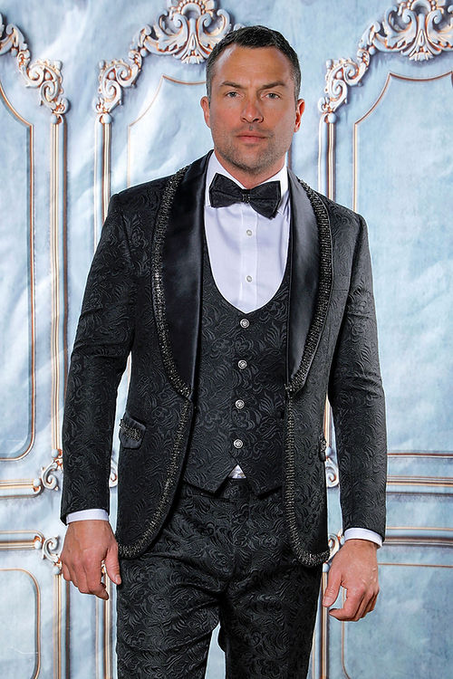 In a grand setting, a man dons the Insomnia MANZINI SOFIA 3-Piece Shiny Rhinestone Trim Floral Textured Black Tuxedo Suit. The ensemble features a tailored fit, bow tie, and satin lapel for added elegance. It's dry clean only to ensure its sophisticated look remains pristine.