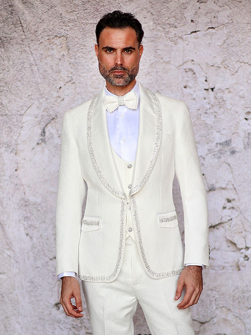 A man stands against a textured light background wearing the Insomnia MANZINI SOFIA 3-piece cream tuxedo suit, featuring a slim fit with satin shawl lapel and bow tie, and adorned with shiny rhinestone trim on the floral texture.