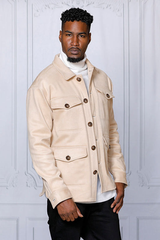 A man in a MANZINI SPAIN Tan Large Pockets & Button Winter Coat by Insomnia, layered over a white turtleneck sweater, stands elegantly against a light gray ornate background.