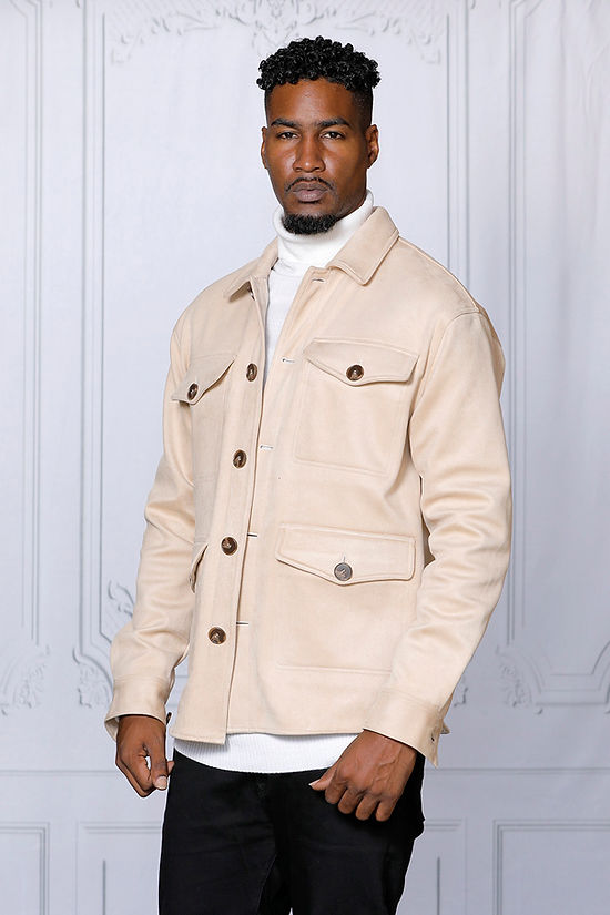 A man in a MANZINI SPAIN Tan Large Pockets & Button Winter Coat by Insomnia, layered over a white turtleneck sweater, stands elegantly against a light gray ornate background.