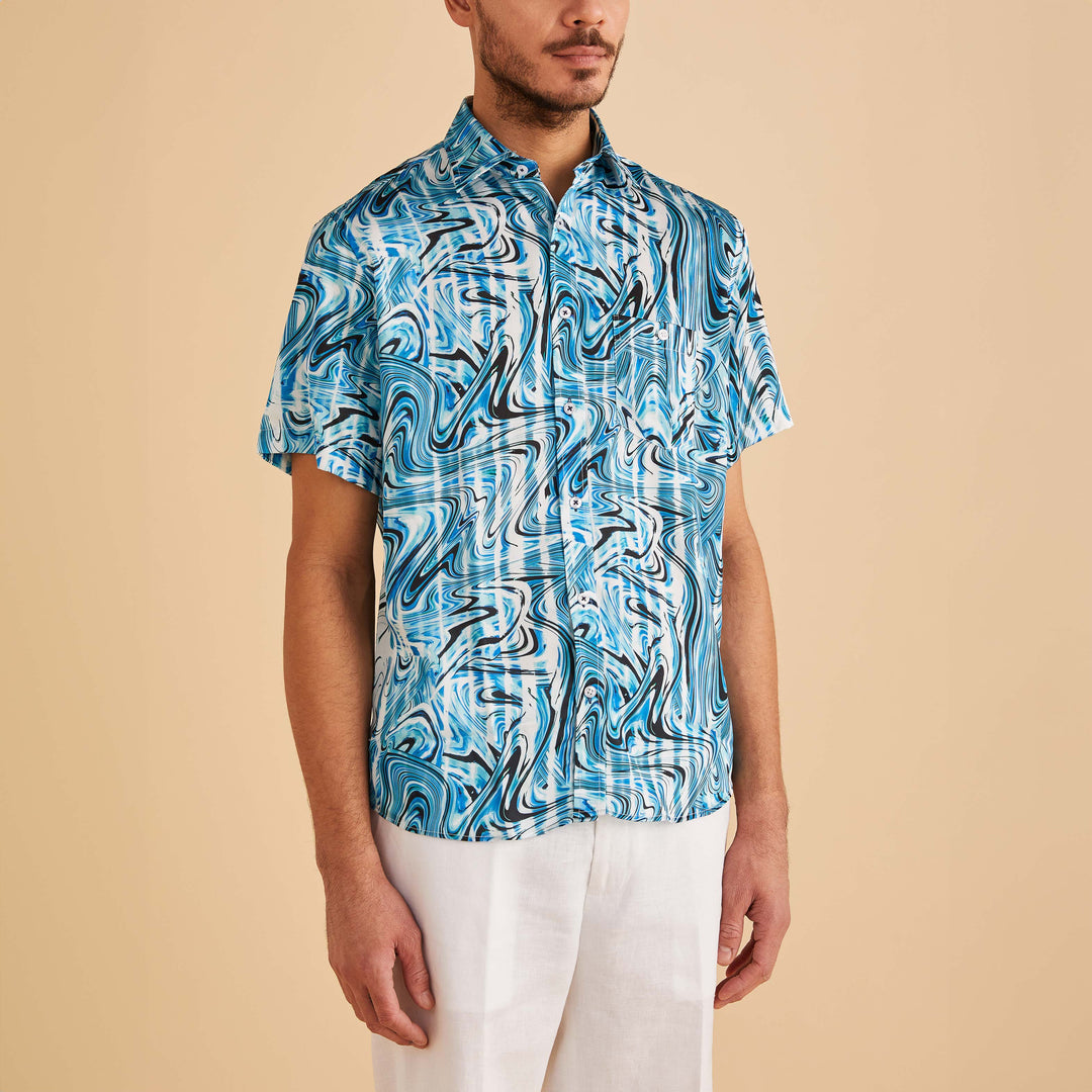 A man wears the Inserch SS Swirl Print Shirt SS012-00195 Indigo, featuring blue and white wave designs, paired with white pants, as he stands against a plain beige background.