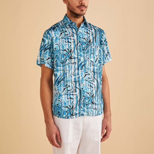 A man wears the Inserch SS Swirl Print Shirt SS012-00195 Indigo, featuring blue and white wave designs, paired with white pants, as he stands against a plain beige background.