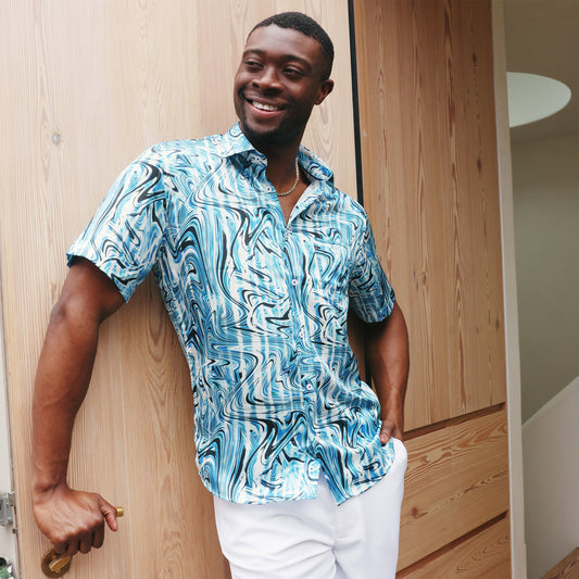 A man wears the Inserch SS Swirl Print Shirt SS012-00195 Indigo, featuring blue and white wave designs, paired with white pants, as he stands against a plain beige background.