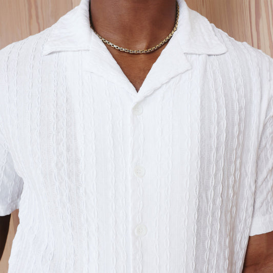 Someone wearing the Inserch SS Knit Camp Collar Shirt SS020-00002 White by Inserch and a gold chain necklace stands in front of a light wood background.