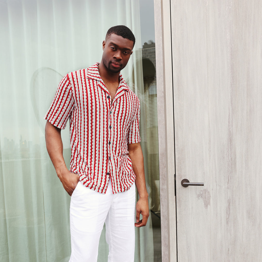 A man wearing the Inserch SS Knit Camp Collar Shirt SS020-00030 Red by Inserch, featuring red, white, and black vertical stripes, is paired with white pants and stands against a beige background.