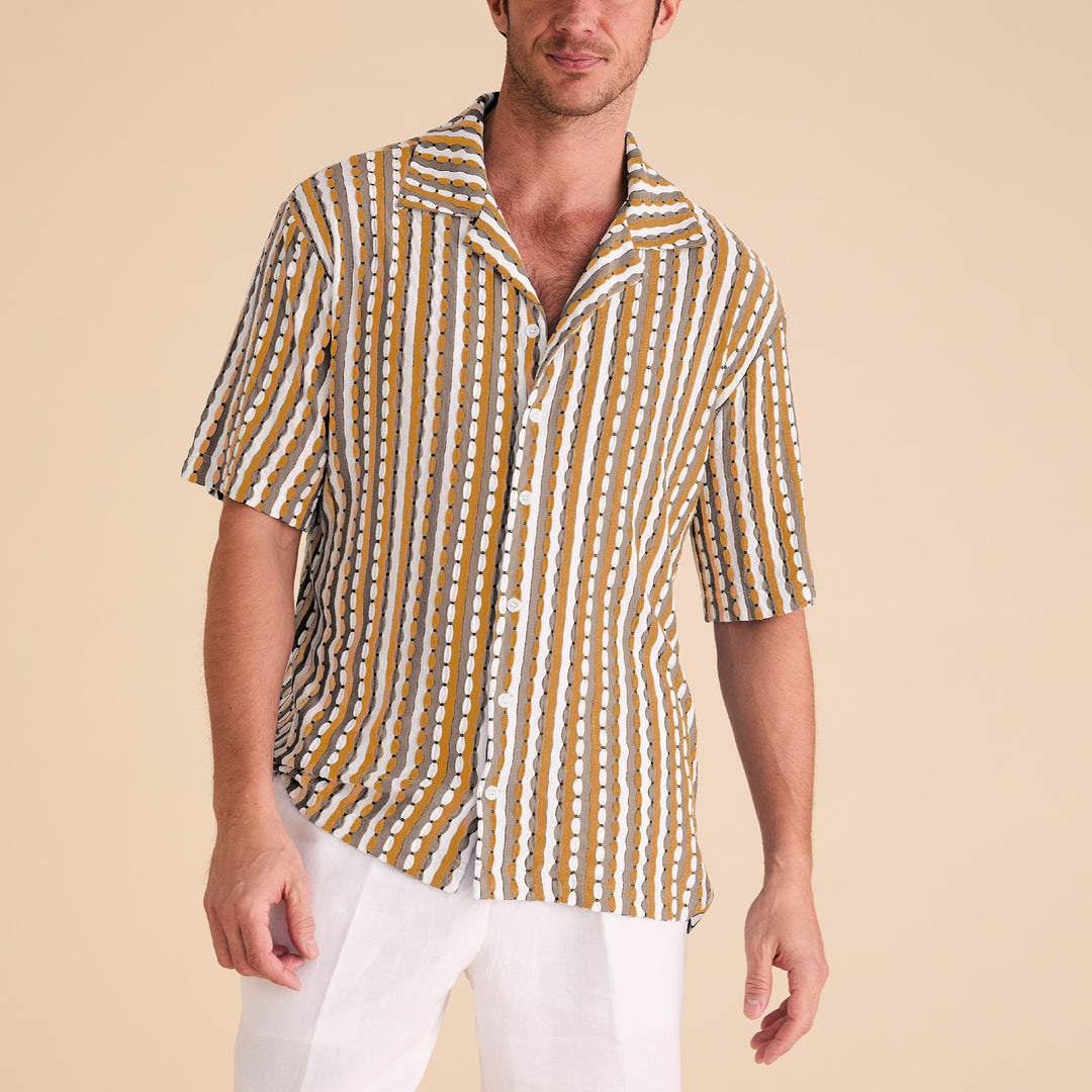 Man wears the Inserch SS Knit Camp Collar Shirt SS020-00038 Gold by Inserch, featuring mustard yellow, white, and gray vertical stripes, paired with white pants against a beige background.