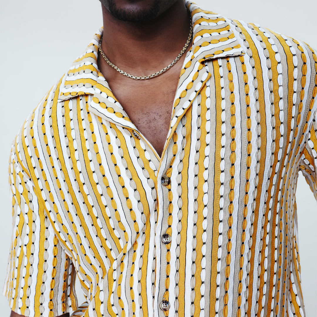 Man wears the Inserch SS Knit Camp Collar Shirt SS020-00038 Gold by Inserch, featuring mustard yellow, white, and gray vertical stripes, paired with white pants against a beige background.