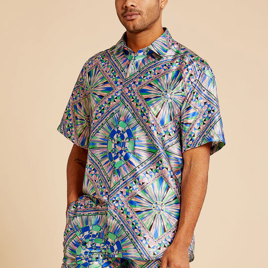 A man wears the Inserch SS Kaleidoscope Print Shirt SS052-00126 Purple by Inserch, featuring a vibrant geometric satin pattern in blue, green, and purple, standing with one hand in his pocket against a beige background.