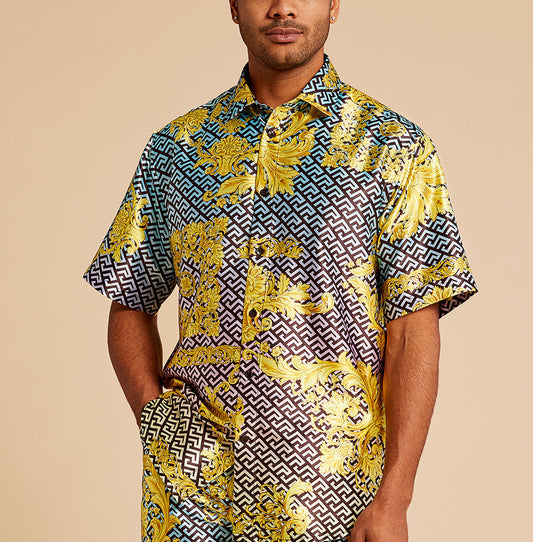 A man wears the Inserch SS Grecian Print Shirt SS053-00066 Multi—a short-sleeve shirt with bold geometric and baroque yellow patterns—standing against a plain beige background.