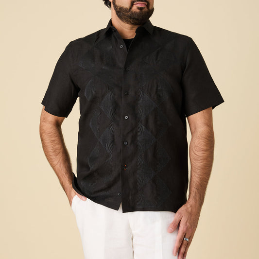 A man wearing the Inserch SS Linen Embroidered Shirt SS126-00001 in black pairs it with white shorts against a beige background.