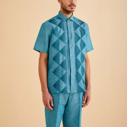 A man in an Inserch SS Linen Embroidered Shirt SS126-00172 Caspian, a teal short-sleeve button-up made from premium European linen, paired with matching geometric pants, stands against a plain beige background.