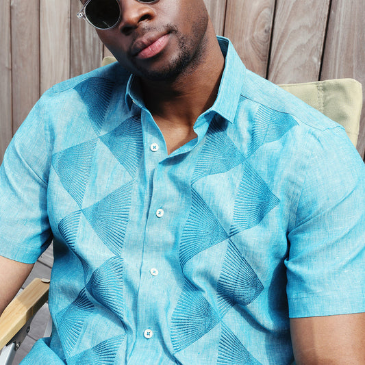 A man in an Inserch SS Linen Embroidered Shirt SS126-00172 Caspian, a teal short-sleeve button-up made from premium European linen, paired with matching geometric pants, stands against a plain beige background.