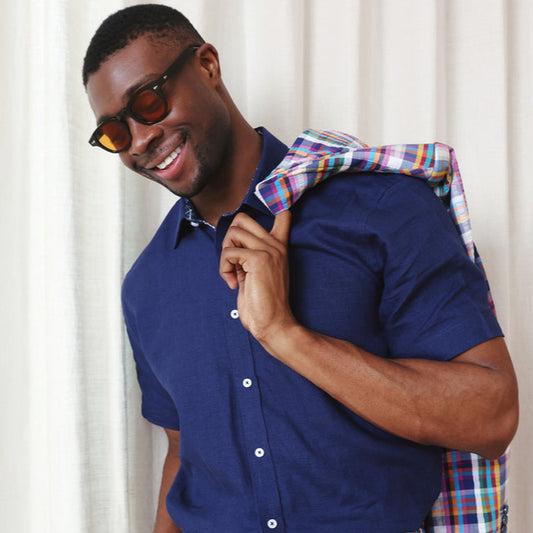A man in the Inserch Linen 2pc Set (SS717) Ink—a navy short-sleeve shirt and multicolored plaid pants—holds a matching jacket over his shoulder, standing before a white curtain.