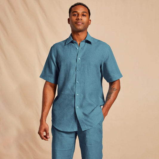 A person wearing the Inserch SS Linen Shirt SS717-00172 Caspian, a blue short-sleeve button-up with floral print trim, along with premium linen pants stands against a beige background.