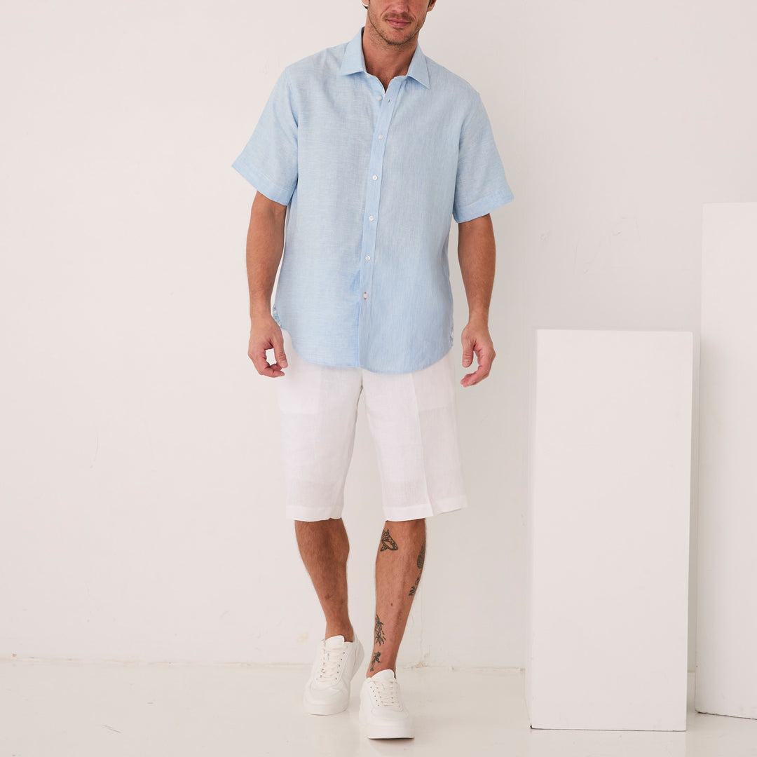 A man in an Inserch SS Linen Shirt SS717-00183 Blue Mist and white shorts stands indoors near white blocks, wearing white sneakers.