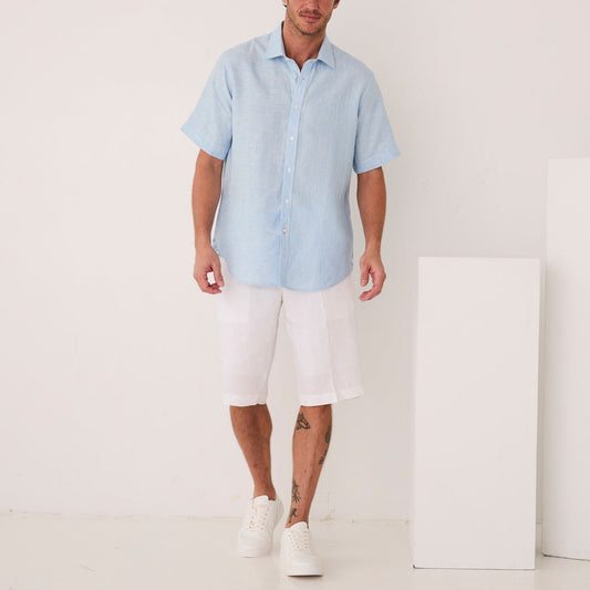 A man in an Inserch SS Linen Shirt SS717-00183 Blue Mist and white shorts stands indoors near white blocks, wearing white sneakers.