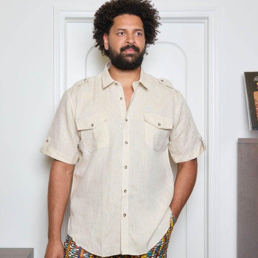 A man stands indoors, facing forward with one hand in his pocket, wearing the Inserch SS Linen Military Shirt (SS718-00006 Oatmeal), a light beige short-sleeve shirt with two chest pockets by Inserch, paired with patterned pants.