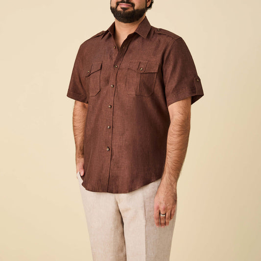 A man wears the Inserch SS Linen Military Shirt SS718-00024 in Chocolate by Inserch, featuring short sleeves and two chest pockets, paired with light-colored pants against a beige background.
