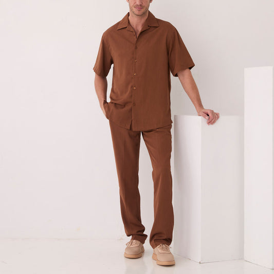 A man stands indoors in the Inserch Crepe Camp Collar Shirt SS9356-00064 Mocha by Inserch, paired with matching brown pants and beige shoes, one hand in his pocket and the other resting on a white structure.