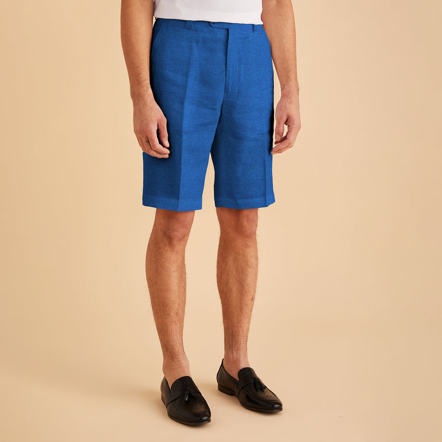 A person wearing Inserch Modern Linen Shorts in River Blue, paired with a crisp white shirt and black loafers, stands against a beige background.