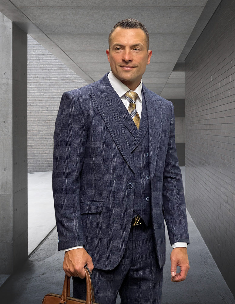 A man in a STATEMENT CLOTHING | STEFANO-NAVY checked Italian wool suit by Statement Clothing holds a brown bag, standing in a sleek concrete and brick corridor.