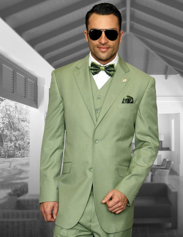 A man with dark hair and sunglasses poses indoors wearing the STATEMENT CLOTHING | STZV-102-APPLEGREEN modern fit suit, matching vest, and bow tie by Statement Clothing, with one hand slightly raised.
