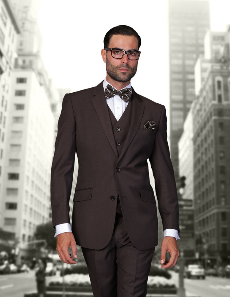 A man wearing the STATEMENT CLOTHING | STZV-102-BROWN modern fit suit by Statement Clothing, with a bow tie and glasses, stands before a blurred city street with tall buildings.