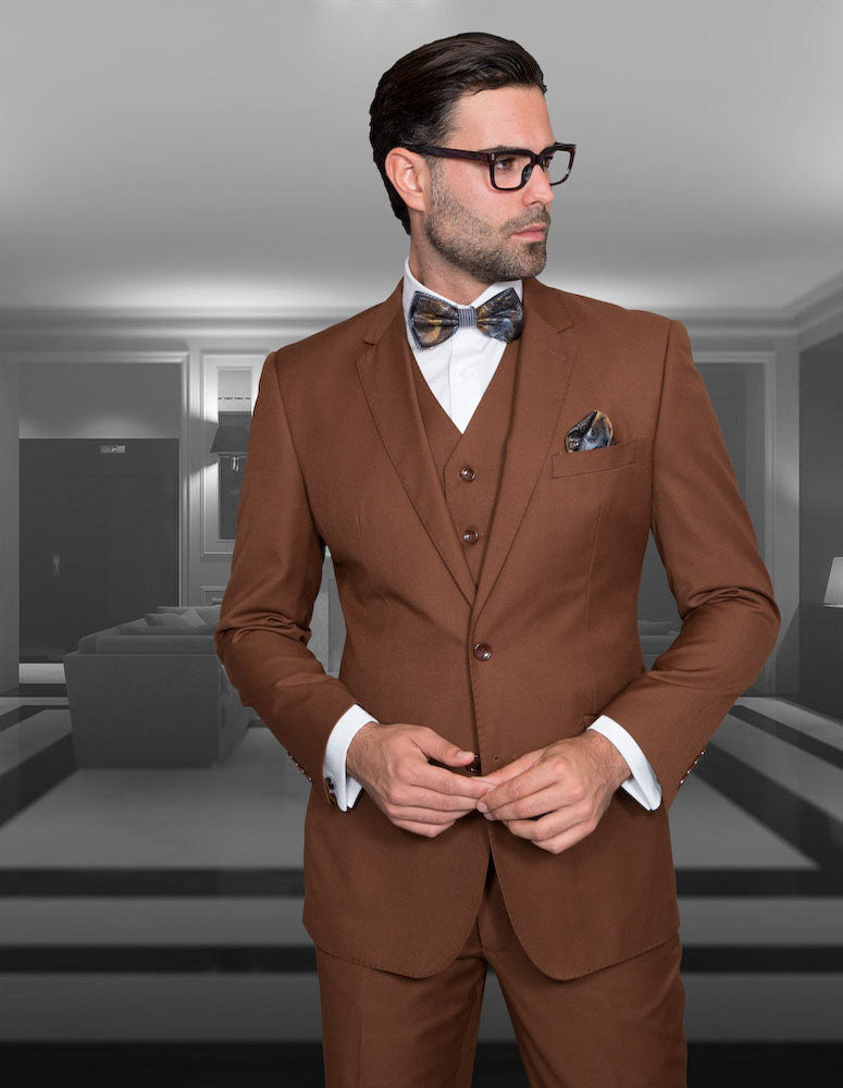 A man in the STATEMENT CLOTHING | STZV-102-COPPER suit by Statement Clothing, with a bow tie and glasses, stands before a modern, monochrome living room.