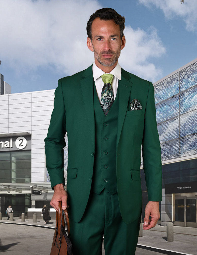 A man in a Statement Clothing STATEMENT CLOTHING | STZV-102-FORESTGREEN Italian wool suit holds a brown briefcase outside an airport terminal under a partly cloudy sky.