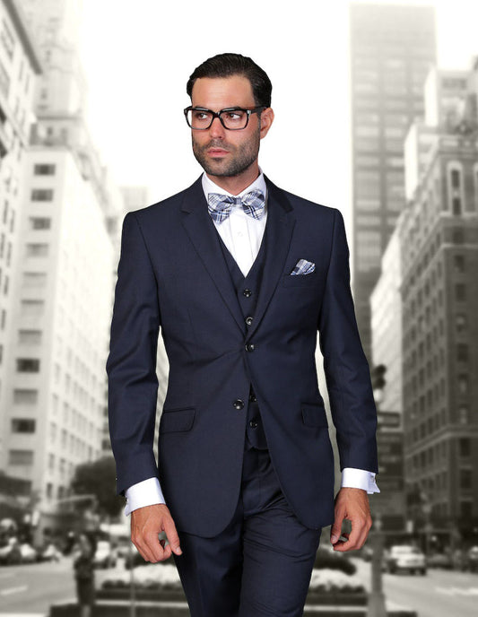 A man in the STATEMENT CLOTHING | STZV-102-NAVY modern fit navy suit by Statement Clothing, paired with a bow tie and glasses, walks confidently on a city street lined with tall buildings.