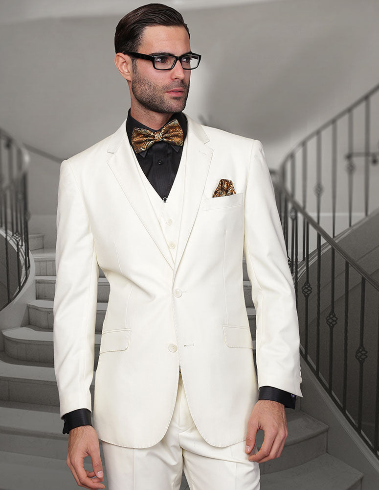 A man in the STATEMENT CLOTHING | STZV-102-OFFWHITE modern fit suit from Statement Clothing, paired with a black shirt, gold patterned bow tie, and matching pocket square, stands before a staircase.