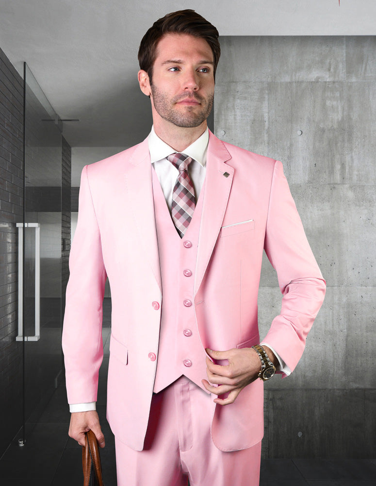 A man in a modern fit, light pink STATEMENT CLOTHING STZV-102-PINK Italian wool suit by Statement Clothing stands indoors with a plaid tie and brown bag, set against concrete and glass walls.