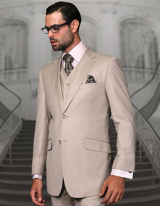 A man wearing the STATEMENT CLOTHING | STZV-102-TAN, a modern fit light beige Italian wool suit by Statement Clothing with vest, patterned tie, and pocket square stands before an ornate staircase, gazing to the side.