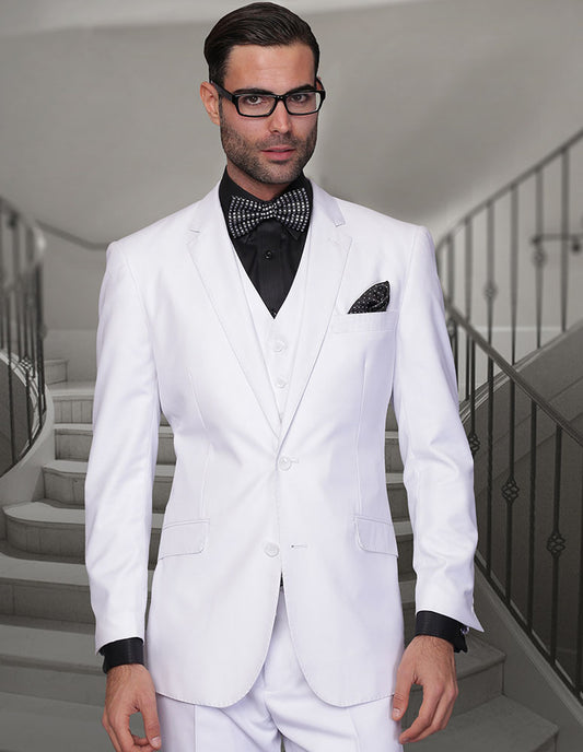 A man in the STATEMENT CLOTHING | STZV-102-WHITE modern fit Italian wool suit by Statement Clothing, paired with a black shirt, black bow tie with white dots, and glasses, stands in front of a staircase.