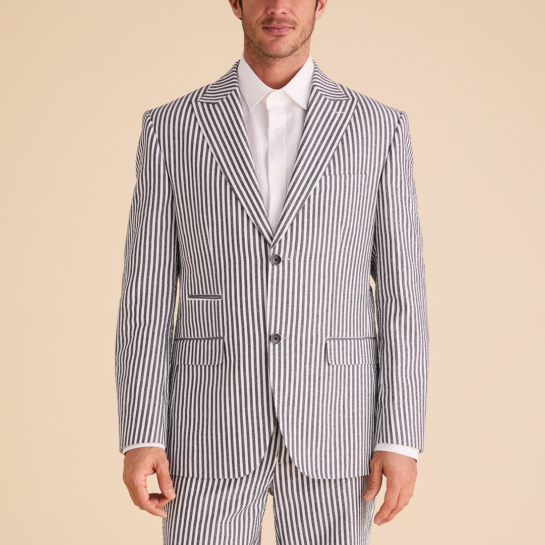 A man dons the Inserch Seersucker Stripe Blazer SU660155-00001 Black, paired with a white shirt, standing against a beige background.