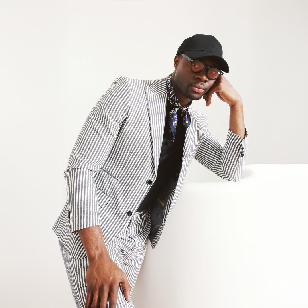 A man dons the Inserch Seersucker Stripe Blazer SU660155-00001 Black, paired with a white shirt, standing against a beige background.