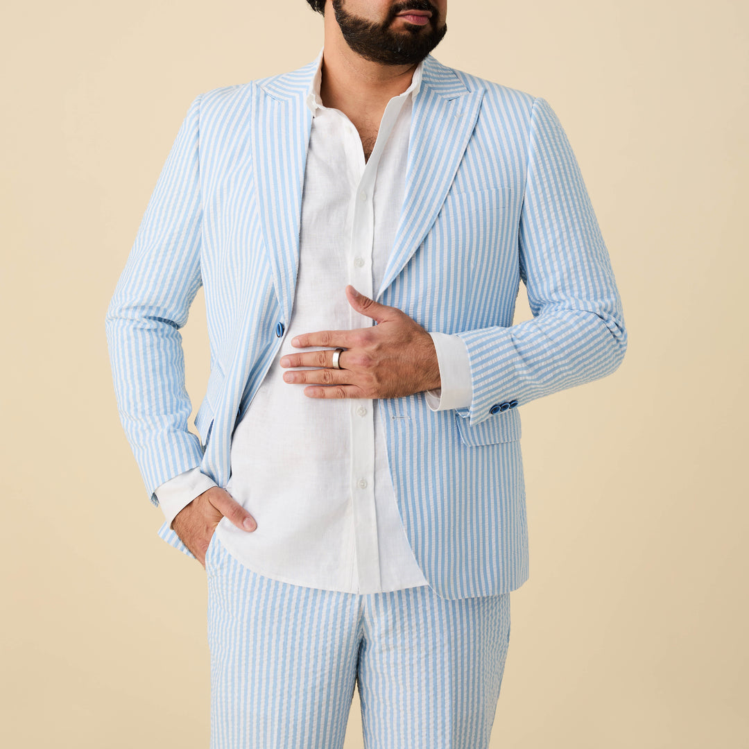 A man wears the Inserch Seersucker Stripe Blazer SU660155-00014 in light blue with a white shirt, standing against a beige background. His left hand rests on his stomach while his right is in his pocket. Brand: Inserch.