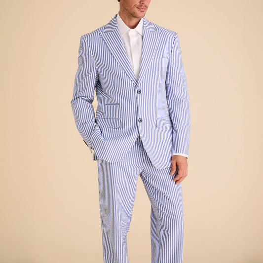 A person confidently stands with hands in pockets, dressed in Inserch Seersucker Stripe Pants P660155-00101 True Blue and a blue and white pinstripe suit with a modern fit, paired with a crisp white shirt and brown leather dress shoes.