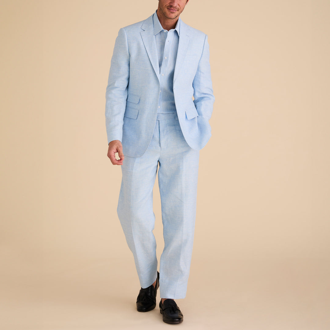 Showcasing the Inserch Slim Linen Suit SU880-00183 in Blue Mist, a person stands poised against a beige background, pairing the serene set with matching shirts and sleek black shoes. This ensemble underscores the sophisticated texture of its yarn-dye fabric.