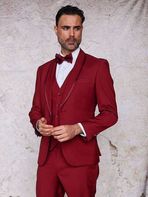A man in an Insomnia MANZINI SUNSET slim fit burgundy tuxedo with a reversible vest and a bow tie stands against a textured beige background.