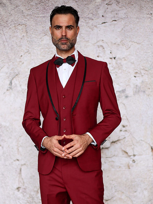 A man in an Insomnia MANZINI SUNSET Slim Fit 3-Piece Burgundy Black Tuxedo Suit with a bow tie stands against a textured gray wall, looking forward with hands clasped.