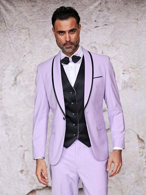 A man dressed in an Insomnia MANZINI SUNSET Lavender Tuxedo Suit with a reversible vest and bow tie stands against a textured wall.
