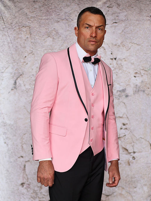 A man poses against a textured backdrop wearing an Insomnia MANZINI SUNSET Slim Fit 3-Piece Pink Black Tuxedo Suit with a reversible vest and bow tie.