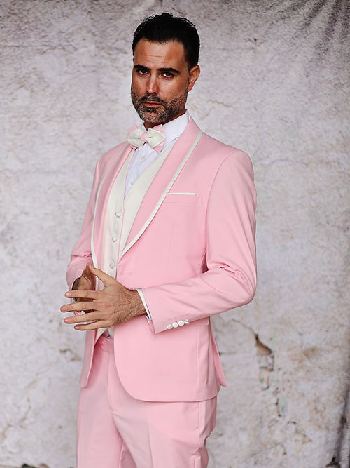 A man confidently poses against a textured wall wearing the Insomnia MANZINI SUNSET Slim Fit 3-Piece Pink White Tuxedo Suit with a reversible vest and white bow tie.