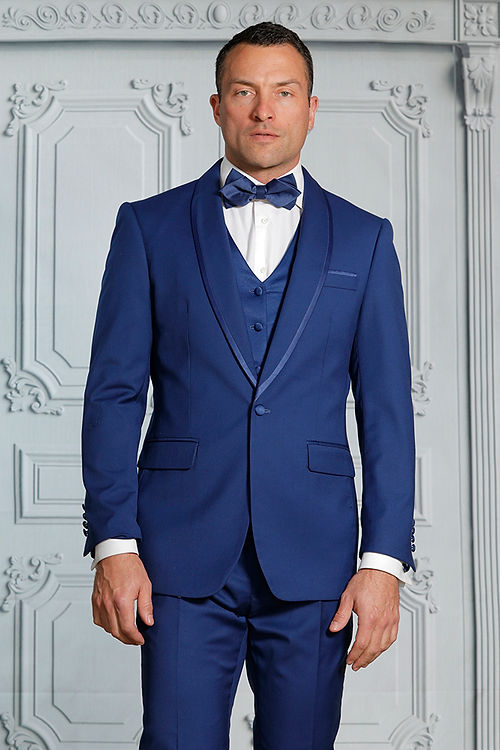 Dressed in the MANZINI SUNSET Slim Fit 3-Piece Sapphire Tuxedo Suit with a reversible vest by Insomnia, a man in a blue suit with a white shirt and bow tie stands before an ornate gray wall, exuding modern elegance.
