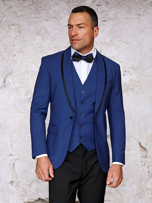 A man wearing the Insomnia MANZINI SUNSET Slim Fit Sapphire Black Tuxedo with a reversible vest and black bow tie stands against a textured wall.