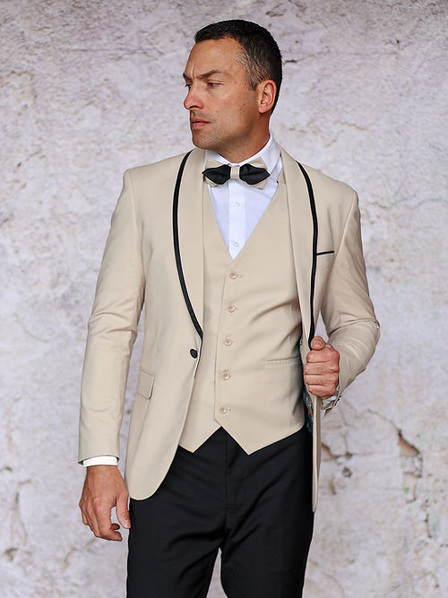 A man adjusts his jacket in an Insomnia MANZINI SUNSET Slim Fit 3-Piece Tan Tuxedo Suit with a reversible vest, complemented by a black bow tie and white shirt. The suit is made from soft, breathable fabric for ultimate comfort.
